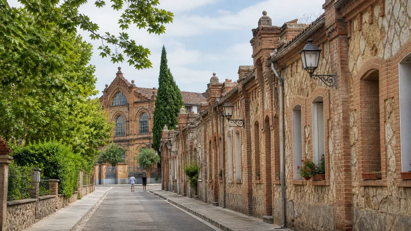 Calle arbolada de la Colònia Güell con viviendas de ladrillo y piedra y un equipamiento al fondo