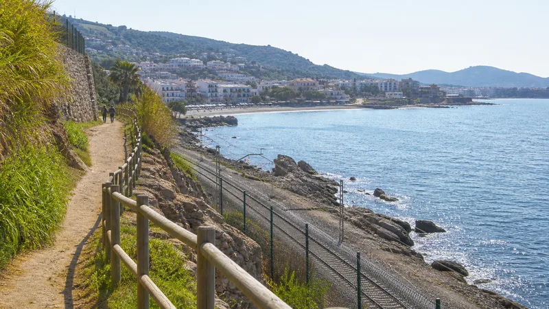Camino litoral protegido sobre roca con vía ferroviaria vallada y cala al fondo