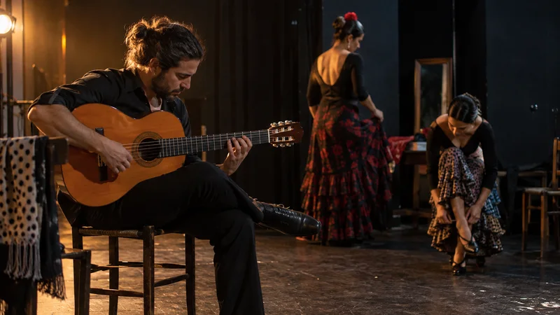 Un guitarrista ensaya junto a dos bailaoras en un escenario pequeño, con iluminación cálida y fondo oscuro.